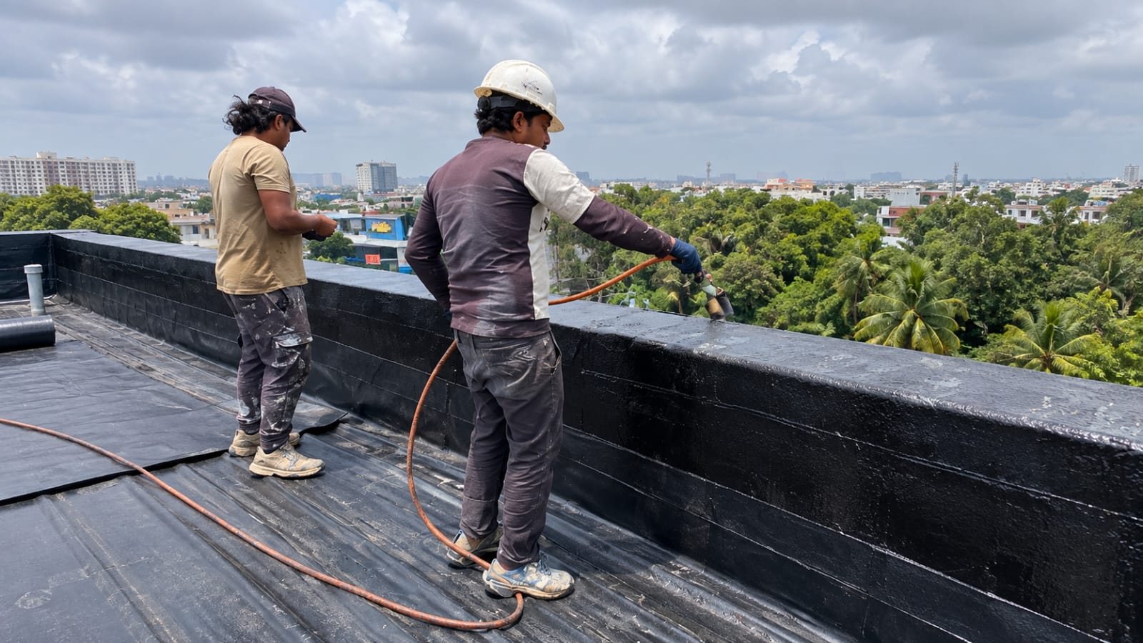 Two workers spraying black waterproof coating onto the outer face of a rooftop parapet wall